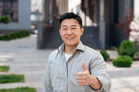 Cheerful middle-aged Asian man giving a thumbs-up outside a modern building, conveying satisfaction and trust in dental care.