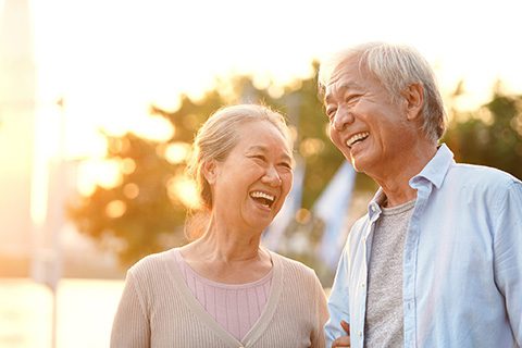 Happy senior Asian couple smiling and enjoying time outdoors at sunset, representing healthy aging and positive dental outcomes.