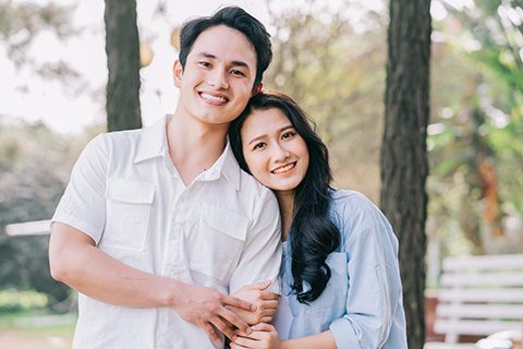 Young Asian couple embracing and smiling in a park, reflecting happiness, healthy relationships, and confident smiles.