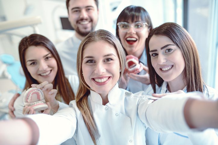Group of smiling dental professionals taking a selfie in a bright, modern clinic, with two team members holding dental models showcasing oral anatomy.